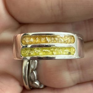 Peut inclure: Une bague en argent avec deux incrustations rectangulaires parallèles. L'incrustation supérieure est dorée et l'incrustation inférieure est de couleur vert clair. La bague est présentée en gros plan, avec une deuxième bague visible en arrière-plan.