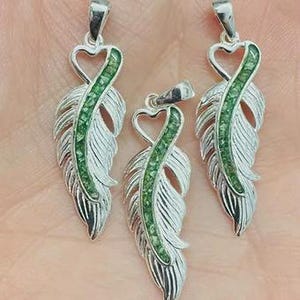 Peut inclure: Trois pendentifs en argent en forme de plume avec un fermoir en forme de cœur. Chaque pendentif présente un centre vert orné de pierres précieuses et un motif de plume détaillé. Les pendentifs sont présentés sur une surface claire.