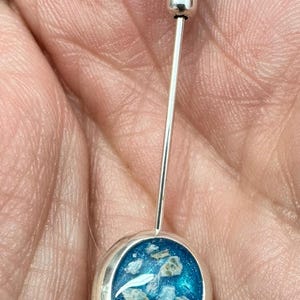 Peut inclure: Une épingle en argent avec un cabochon rond en résine bleue contenant des paillettes blanches et brunes. L'épingle est fixée à une longue tige en argent.