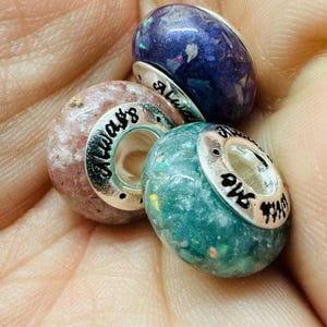 May include: Three glass beads with silver accents and inscribed text. The beads are purple, pink, and aqua  with flecks of glitter. The text on the beads reads "Always", "Love", and "And", respectively.