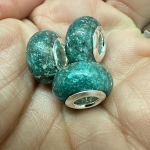 May include: Three teal green glass beads with silver metal accents. The beads have a speckled texture.