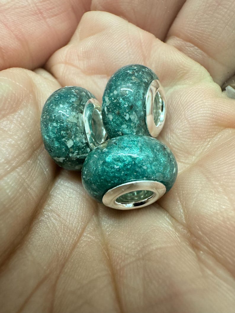 May include: Three teal green glass beads with silver metal accents. The beads have a speckled texture.