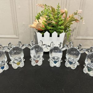 May include: Six clear glass reindeer figurines with a heart-shaped detail in the center of their chest. Each reindeer has a different colored heart, including blue, gold, red, and green.