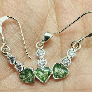 Puede incluir: Un conjunto de joyas de plata con pendientes y un colgante. Cada pieza tiene una piedra verde en forma de corazón y tres piedras redondas y transparentes. Los pendientes tienen un diseño de gancho, y el colgante tiene un pequeño lazo.