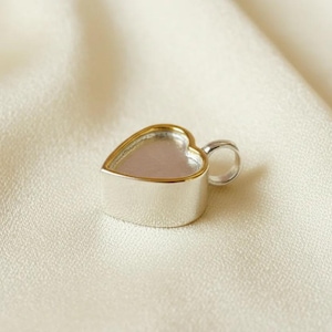 May include: A silver heart-shaped pendant with a gold-colored rim. The pendant has a small loop for a chain and sits on a cream-colored fabric.