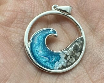 Può includere: Un ciondolo circolare in argento con un'onda blu e sabbia bianca all'interno. L'onda è realizzata in resina e la sabbia è vera.