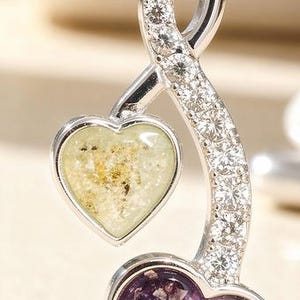 Peut inclure: Pendentif argenté avec deux breloques en forme de cœur. L'un des cœurs est rempli d'une matière tachetée vert clair, l'autre d'une matière violette. La tige incurvée du pendentif est incrustée de petits cristaux transparents.