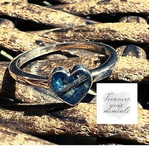 Peut inclure: Une bague en argent avec une pierre bleue et noire en forme de cœur. La bague est sur un fond brun tissé. Le texte "Treasure your moments" est dans le coin inférieur droit de l'image.