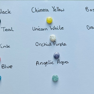May include: A white card with 10 small round glass beads in different colors, each with a handwritten label describing the color. The colors are: Vampire Black, Chimera Yellow, Basilisk Green, Mermaid Teal, Unicorn White, Dragon Blood Red, Fairy Pink, Orchid Purple, Aegean Blue, and Angelic Aqua.
