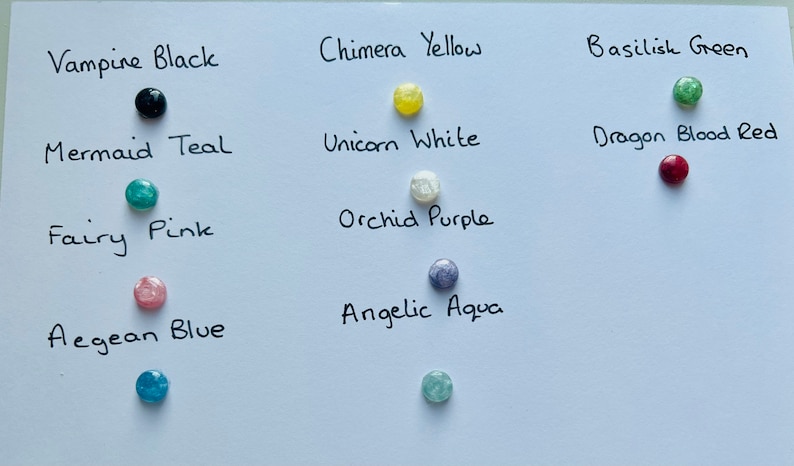 May include: A white card with 10 small round glass beads in different colors, each with a handwritten label describing the color. The colors are: Vampire Black, Chimera Yellow, Basilisk Green, Mermaid Teal, Unicorn White, Dragon Blood Red, Fairy Pink, Orchid Purple, Aegean Blue, and Angelic Aqua.