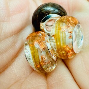 May include: Three glass beads with silver accents. The beads are clear with gold and copper flecks and a brown and black bead.
