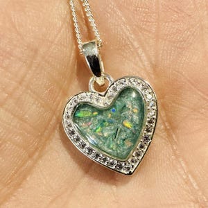 Peut inclure: Un pendentif en argent en forme de cœur avec un insert en résine transparente rempli de paillettes vertes et bleues irisées. Le pendentif est entouré d'une rangée de petites pierres de zircone cubique transparentes.