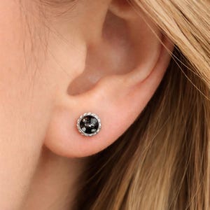 May include: A small, round stud earring with a black and silver speckled design. The earring is set in a silver-coloured metal with a decorative, beaded edge. The earring is shown on an ear.