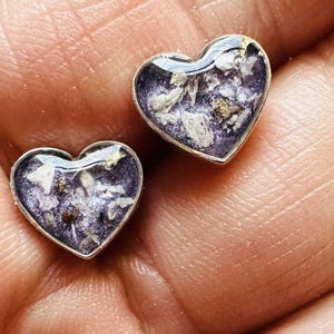 Peut inclure: Une paire de boucles d'oreilles en forme de cœur avec un sertissage argenté. Chaque boucle d'oreille présente une résine transparente remplie de fleurs violettes séchées et de points blancs.