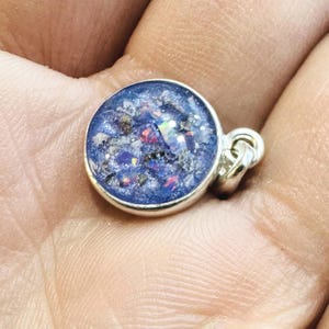 May include: A round, silver pendant with a blue resin center. The resin contains iridescent flecks of color. The pendant is held in a hand, showcasing its size and design. The pendant has a small loop for a chain.