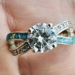Peut inclure: Une bague en argent avec une grosse pierre transparente de taille ronde en son centre. La monture comporte de petites pierres et des incrustations de couleur turquoise, créant un design unique et élégant. La bague est présentée en gros plan.