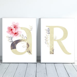 May include: Two white framed prints with gold letters, "A" and "R", decorated with pink watercolor flowers and gold leaf accents.