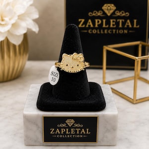 May include: A gold-toned ring featuring a textured Hello Kitty face design with a bow on the side. A small white circular tag attached to the band displays the text SIZE 10 in black font. The ring is displayed on a black velvet cone stand.