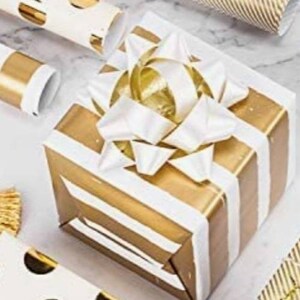 Könnte beinhalten: Eine weiße und gold gestreifte Geschenkbox mit einer großen weißen Schleife. Die Schleife ist mit einem goldenen Band gebunden.