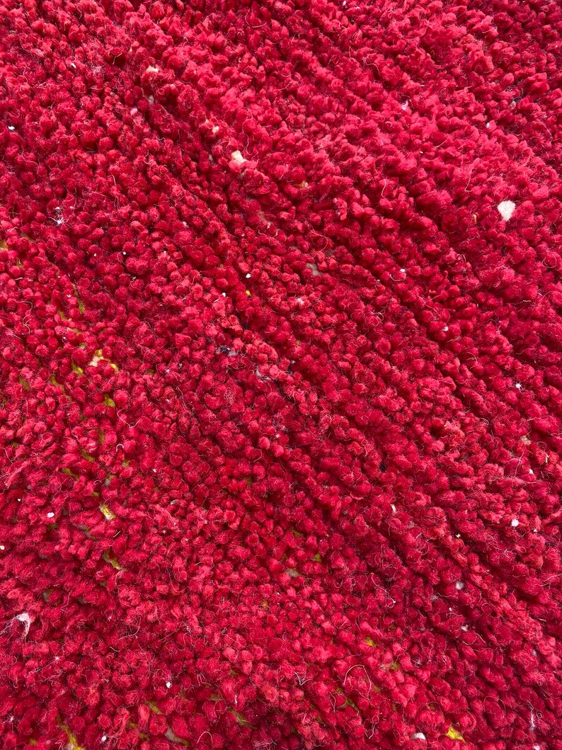 May include: Close-up of a vibrant red rug with a textured, plush surface. The rug's color is a deep, rich red, with a slightly uneven texture. Small flecks of white and yellow are visible throughout the rug.