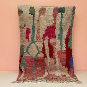 May include: A rectangular, hand-woven rug with a colourful, abstract design. The rug features a beige background with vertical and horizontal shapes in shades of red, pink, blue, and green. The rug has a fringed edge.