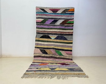 May include: A rectangular, handwoven rug with a striped pattern in various colors. The rug features horizontal stripes in shades of gray, pink, yellow, purple, and green, with geometric shapes. The rug has a fringed edge.