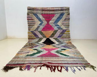 May include: A colorful, geometric rug with a central diamond pattern. The rug features a mix of pink, blue, and neutral tones, with a fringed edge. The rug's design includes chevron and triangle shapes.