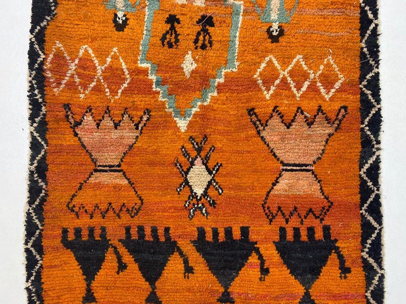 a vibrant, intricately designed rug with a striking orange and black color scheme. The rug is adorned with various geometric patterns and shapes, including triangles, diamonds, and other abstract designs. The rug appears to be a traditional or cultural textile, showcasing the craftsmanship and artistry of the creator.