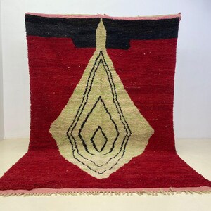 May include: A red and black geometric rug with a central diamond design in beige and black. The rug has a textured surface and a fringed edge. The rug is a decorative floor covering.