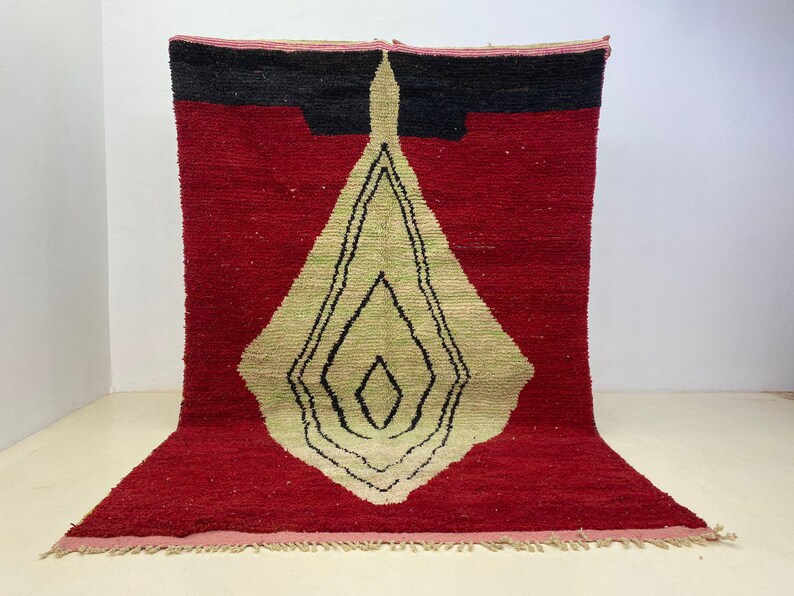 May include: A red and black geometric rug with a central diamond design in beige and black. The rug has a textured surface and a fringed edge. The rug is a decorative floor covering.
