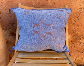 May include: A blue cushion with a brown geometric pattern, placed on a wooden chair. The cushion has tassels on the corners. The background is a textured, orange-brown wall.