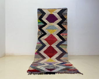 May include: A colorful, geometric patterned rug with a diamond design. The rug features a repeating pattern of diamonds in red, purple, yellow, and blue, outlined in black. The rug is made of woven fabric and has a fringed edge.