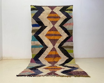 May include: A colorful, geometric rug with a diamond pattern. The rug features a cream-colored background with black, orange, purple, blue, green, and pink zigzags and diamonds. The rug is made of a woven material.