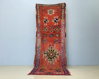 Op de afbeelding: Een rechthoekig, handgeweven tapijt met een rood-oranje achtergrond en geometrische patronen in blauw, groen, geel en zwart. Het tapijt heeft ruit- en stermotieven, met een blauwe en roze rand. Het tapijt wordt verticaal weergegeven.