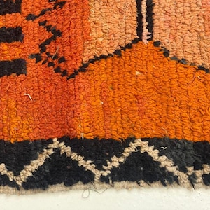 The image is a close-up of a vibrant, multicolored rug with a geometric pattern. The rug has a mix of orange, black, and white colors, creating a visually striking and intricate design.