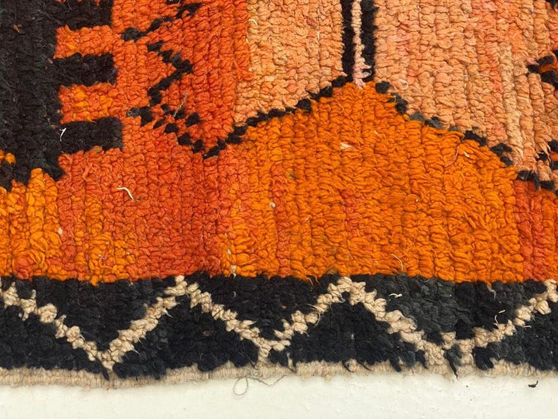 The image is a close-up of a vibrant, multicolored rug with a geometric pattern. The rug has a mix of orange, black, and white colors, creating a visually striking and intricate design.