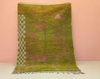 May include: A rectangular, olive green rug with a geometric design in pink. The rug has a checkered border on the left side and a fringe along the bottom edge. The rug is made of a thick, textured material.