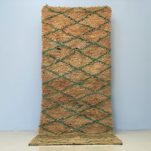 May include: A rectangular rug with a diamond pattern. The rug has a tan background with a teal diamond design. The rug is made of a thick, textured material and is displayed against a light blue wall.