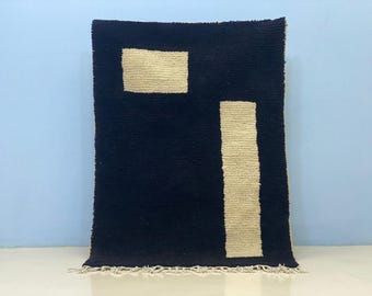 Puede incluir: Alfombra rectangular azul oscuro con un diseño geométrico crema. El diseño incluye un rectángulo y una franja vertical. La alfombra tiene un borde con flecos y se muestra sobre un fondo azul claro. Medidas en centímetros.
