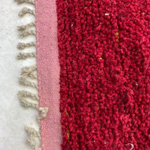 May include: Close-up of a red rug with a textured, plush surface. A light pink border runs along the left side, with off-white fringe detailing. The rug's vibrant color and texture create a cozy aesthetic.