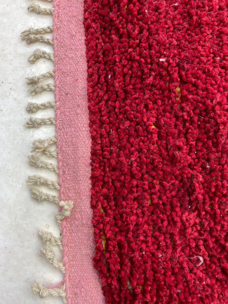 May include: Close-up of a red rug with a textured, plush surface. A light pink border runs along the left side, with off-white fringe detailing. The rug's vibrant color and texture create a cozy aesthetic.