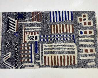 Puede incluir: Alfombra rectangular con fondo gris y patrón geométrico en blanco, marrón y azul. El diseño incluye rayas, cuadrados y formas abstractas, creando una apariencia texturizada. La alfombra probablemente está hecha de un material suave.
