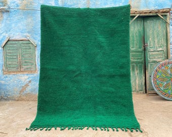May include: A vibrant green rug with a textured surface and a fringe along the bottom edge. The rug is displayed outdoors, against a weathered blue wall and a green door. The rug's color is a rich, solid green.