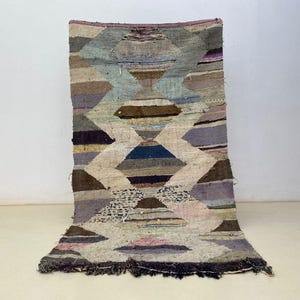 Peut inclure: Un tapis rectangulaire tissé à la main avec un motif géométrique en losange. Le tapis présente une palette de tons neutres, dont le beige, le marron et le gris, avec des touches de bleu, de violet et de rose. Les bords du tapis sont finis avec une frange foncée.