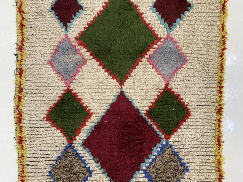 a vibrant, colorful rug with a diamond pattern. The rug is composed of various geometric shapes and colors, including shades of green, red, and yellow. The rug appears to be a traditional, handmade textile, with a textured surface that adds depth and dimension to the design.