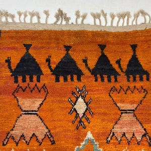 a vibrant, hand-woven rug with a striking geometric pattern in shades of orange and black. The rug is adorned with a variety of shapes and designs, including triangles, diamonds, and other abstract patterns. The rug is framed by a border with a fringe, adding to its overall aesthetic appeal.