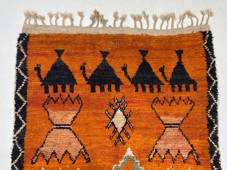 a vibrant, hand-woven rug with a striking geometric pattern in shades of orange and black. The rug is adorned with a variety of shapes and designs, including triangles, diamonds, and other abstract patterns. The rug is framed by a border with a fringe, adding to its overall aesthetic appeal.