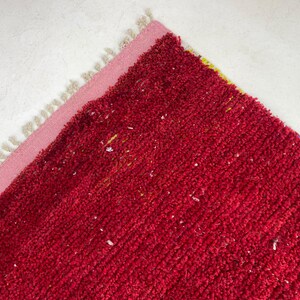 May include: A section of a red rug with a pink border and cream-colored fringe. The rug has a textured surface with small white and yellow flecks. The rug is placed on a white surface.