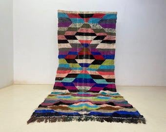 May include: A colorful, geometric patterned rug with a diamond design. The rug features a variety of colors including blue, green, pink, black, and purple. The rug has a fringed edge and is made of woven material.