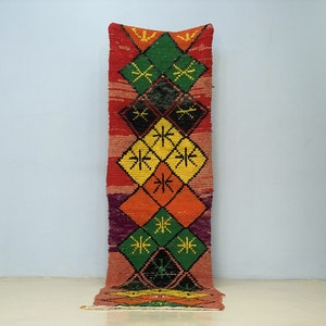 Peut inclure: Un tapis rectangulaire coloré avec un motif de losanges. Le tapis présente un mélange vibrant de teintes rouges, oranges, jaunes, vertes et violettes. Le motif comprend des formes de losanges avec des motifs en forme d'étoiles.